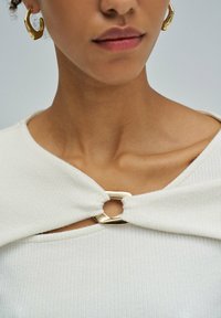 Primo piano del collo e delle labbra di una donna che indossa orecchini a cerchio dorati e un top bianco a coste con una chiusura dorata all'collo.