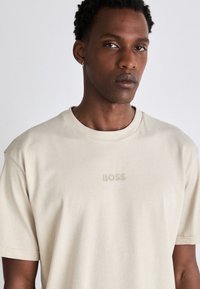 Beige bomull t-skjorte med rund hals og korte ermer. Prentet "BOSS" logo i matchende farge på brystet. Glatt tekstur.