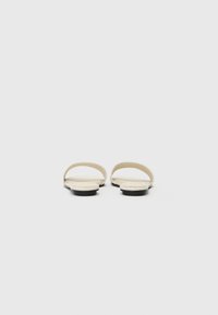 Sandales plates beiges avec deux larges sangles et un design minimaliste. Les semelles noires mettent en valeur la tige claire. Texture lisse sans motifs visibles.
