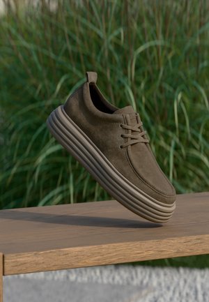 Καφέ suede sneaker με χοντρή, αυλακωτή σόλα, αιωρούμενο πάνω από ξύλινο παγκάκι σε εξωτερικό χώρο με φόντο ψηλό πράσινο χορτάρι.