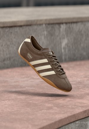 Brun Adidas sneaker med cremede striber og gummisål ser ud til at svæve over en stenoverflade, set fra siden i et udendørs miljø.