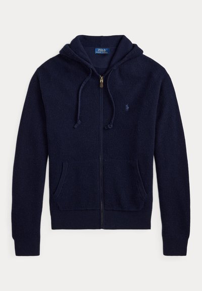 Ζακέτα με κουκούλα σε navy blue, πλεκτή με φερμουάρ, μπροστινές τσέπες, κορδόνι στην κουκούλα και μικρό κεντητό λογότυπο στο στήθος.