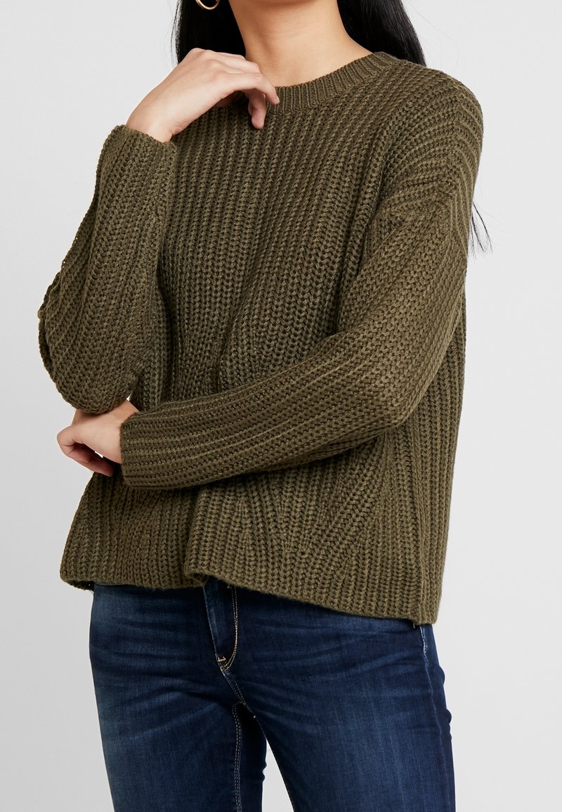 Femme portant un pull en maille grosse olive avec une texture côtelée et un jean bleu foncé, posant avec une main près du visage.
