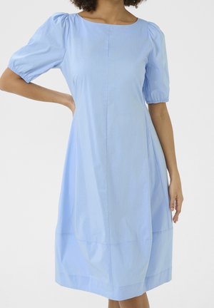 Femme portant une robe bleu clair jusqu'aux genoux avec des manches courtes bouffantes et une taille cintrée, sur un fond uni.