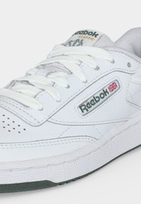 Λευκό παπούτσι Reebok Classic με κορδόνια, διάτρητη μύτη, λογότυπο και σημαία της Ένωσης στη πλαϊνή επιφάνεια.