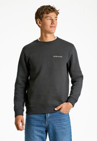 Sudadera de color gris oscuro hecha de una mezcla de algodón, con cuello acanalado, mangas largas y un pequeño logo blanco en el lado izquierdo del pecho. Combinada con jeans azules.