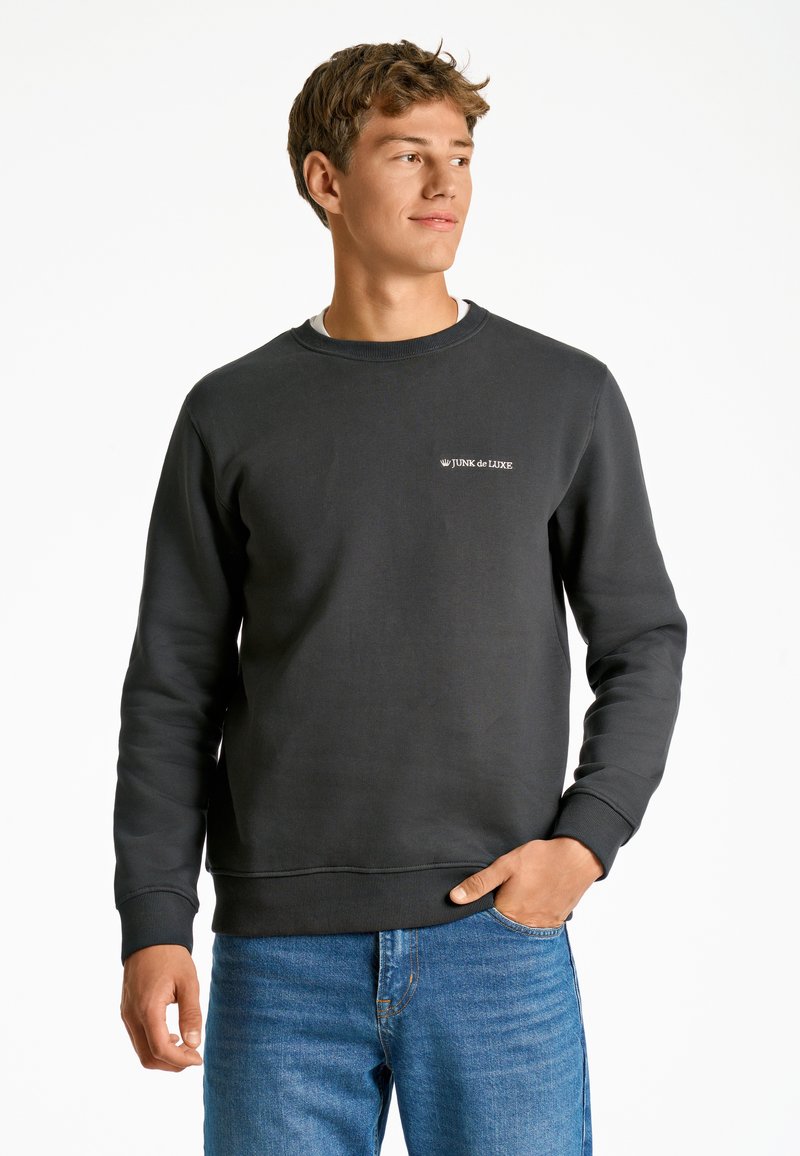 Sudadera de color gris oscuro hecha de una mezcla de algodón, con cuello acanalado, mangas largas y un pequeño logo blanco en el lado izquierdo del pecho. Combinada con jeans azules.