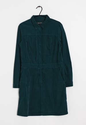 Ciemnozielona sukienka z długim rękawem z sztruksu z kołnierzem, przednie guziki ukryte pod plisą i szwem w talii, wisząca na czarnym wieszaku na białym tle.