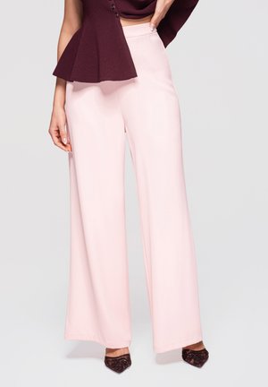 Kvinde iført højtaljede, vidde ben, lyserøde bukser kombineret med mørkelilla pyntede spidse hæle og en matchende mørkelilla top.