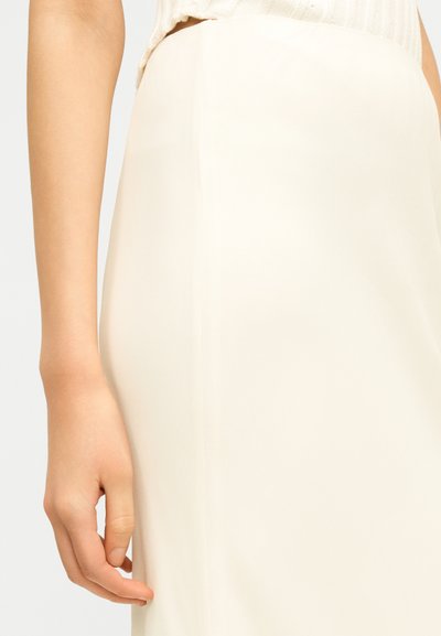 Gros plan sur la main gauche d'une personne reposant à côté d'une jupe couleur crème et d'un haut blanc texturé.