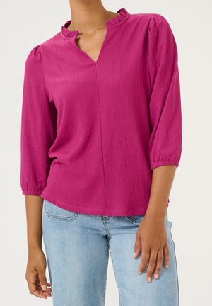 Femme portant un chemisier rose vif à manches longues avec un col à volants et un jean bleu clair sur un fond uni.