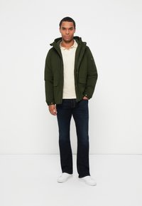 Giacca verde con cappuccio, chiusura con zip, due tasche frontali. Polo di colore beige chiaro, jeans blu scuro e sneaker bianche.