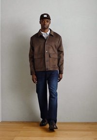 Giacca marrone con bottoni e due tasche frontali, indossata sopra una maglietta a righe e jeans scuri. Scarpe scure completano l'outfit.