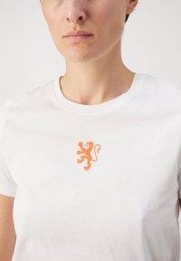 Hvit bomulls t-skjorte med et oransje løvegrafikk på brystet. Designet er enkelt med rund hals og korte ermer.