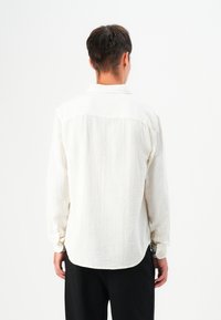 Camisa de manga larga ligera en tejido blanco texturizado con puño abotonado y dobladillo curvado, mostrada desde atrás sobre un fondo liso.