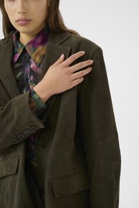 Dunkelgrünes Wildlederblazer mit oversized Passform, Reverskragen, zwei Fronttaschen und schwarzen Knöpfen; getragen über einem bunten gemusterten Hemd.