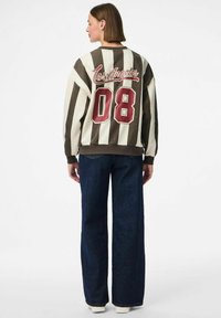 Il pullover presenta strisce marroni e crema, una grafica "Los Angeles", un grande "08" bordeaux e polsini elasticizzati, abbinato a jeans a gamba larga.