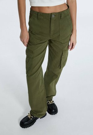 Femme portant un pantalon cargo vert olive avec poches latérales, des mocassins noirs épais avec chaînes dorées, et un top blanc court sans manches.