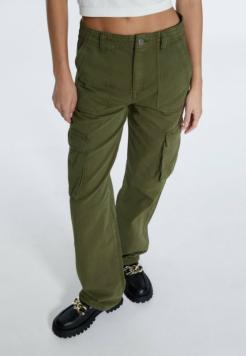 Femme portant un pantalon cargo vert olive avec poches latérales, des mocassins noirs épais avec chaînes dorées, et un top blanc court sans manches.
