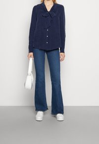 Granatowa bluzka z długim rękawem i kokardą przy dekolcie, zapięcie na guziki z przodu; zestawiona z rozszerzonymi niebieskimi dżinsami i białymi trampkami. Trzyma białą torbę.