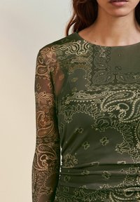 Olīvu zaļš kleita ar garām caurspīdīgām piedurknēm, ar sarežģītiem paisley rakstiem bēšā un brūnā krāsā, kā arī pieguļošu siluetu ar uzziedētu detaļu.