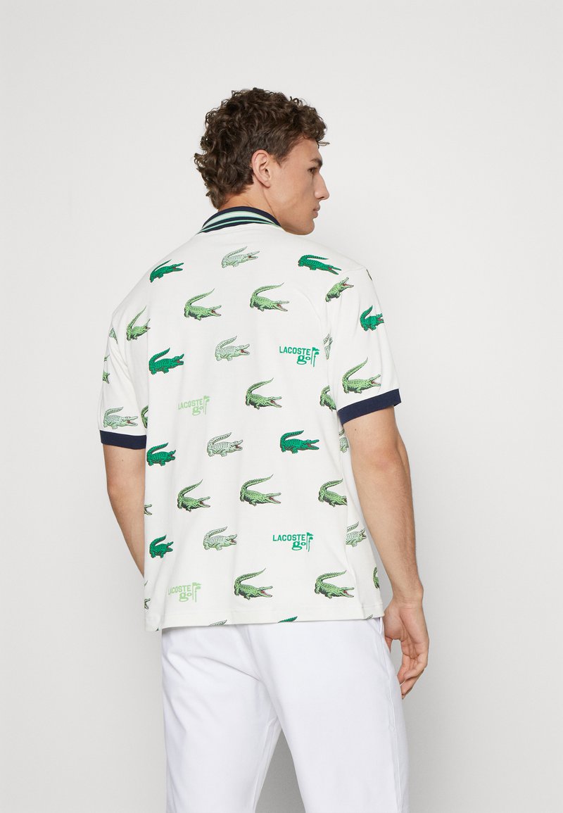 Polo avec un fond blanc présentant des logos de crocodile Lacoste verts. Col et poignets bleu marine, coupe standard, en tissu coton.