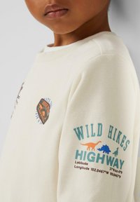 Cremefarbener Sweatshirt mit Dinosauriergrafiken und Text auf den Ärmeln. Hergestellt aus weichem Stoff, mit rundem Ausschnitt und gerippten Bündchen.
