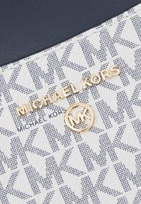 Hvid tekstureret overflade med blåt MK-logo mønster, guld "MICHAEL KORS" tekst og cirkulær gul accent med sammenflettet MK-logo.