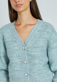 Cardigan bleu deschis cu design în formă de V, țesătură texturată, și cinci butoane decorative, inclusiv trei accente asemănătoare perlelor.