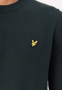 Suéter de punto verde oscuro con cuello redondo y un logotipo de águila bordado en dorado en el lado izquierdo del pecho. Textura suave, diseño ajustado.