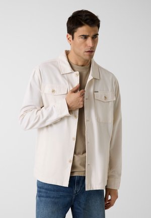 Homme portant une veste beige boutonnée par-dessus une chemise marron et un jean bleu, ajustant le col de la veste, sur un fond clair uni.