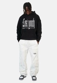 Crna dukserica s bijelim grafičkim tekstom, uparenim s bijelim cargo hlačama koje imaju više džepova i logotip. Tenisice s crno-bijelim naglascima.