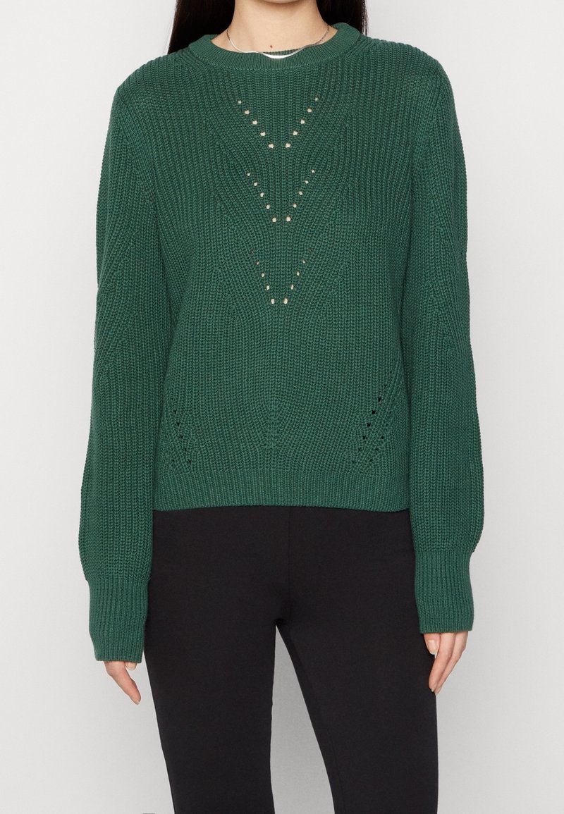 Pull vert tricoté avec un motif en V texturé et de petits détails ajourés, porté avec un pantalon noir ajusté sur une personne debout.