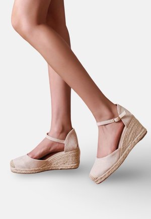 Jambes portant des espadrilles compensées beiges avec des brides à la cheville, présentées de profil sur un fond clair uni.