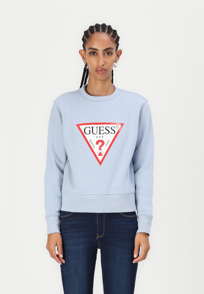Felpa azzurra chiara con colletto e polsini a coste. Presenta un logo triangolare rosso con la scritta "GUESS" e un "?". Indossata con jeans scuri.