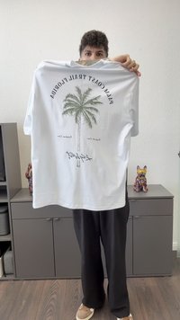 Inimene hoiab valget T-särki, millel on palmipuude graafika ja tekst "Palm Coast Trail Florida", toas, kus on hallid kapid ja dekoratiivsed koerafiguurid.