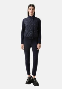 Granatowa pikowana kurtka z zatrzaskami, wysokim kołnierzem i ściągaczami, w połączeniu z dopasowanymi granatowymi spodniami i masywnymi czarnymi butami.