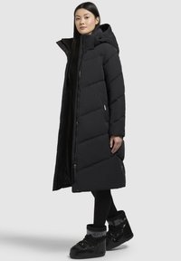 Geacă puffer neagră cu glugă, design lung, model quilted și fermoar frontal. Purtați cu cizme negre izolate.