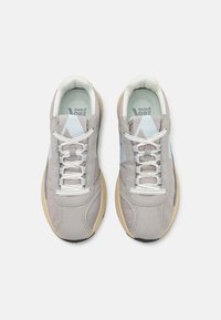 Lys grå sneakers med en teksturert overdel, som har blå detaljer og hvite lisser. Sålen er beige med et mønstret svart bunn.