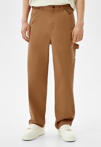 Brązowe spodnie cargo o luźnym kroju, wykonane z lekkiego materiału, z bocznymi kieszeniami i zapięciem na guzik. W połączeniu z białymi trampkami.