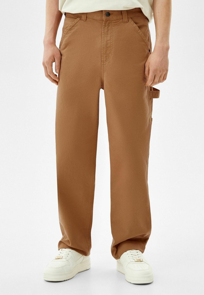Brązowe spodnie cargo o luźnym kroju, wykonane z lekkiego materiału, z bocznymi kieszeniami i zapięciem na guzik. W połączeniu z białymi trampkami.
