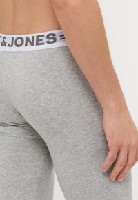 Harmaat legginsit, joissa on valkoinen elastinen vyötärönauha mustalla tekstillä. Kangas näyttää pehmeältä ja siinä on sileä, tasainen pinta.