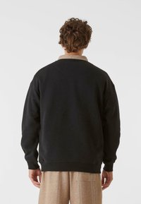 Melnā sporta krekla ar apaļu kaklu un pārmērīgu griezumu, izgatavots no mīksta materiāla ar kontrastējošu beigu apkakli, uzvelkts virs tekstūrētām biksēm.