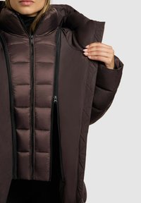 Brun pufferjakke med høj krave, der har et quiltet design, dobbelte lynlåse og en glat overflade. Tydelige teksturerede polstrede paneler.