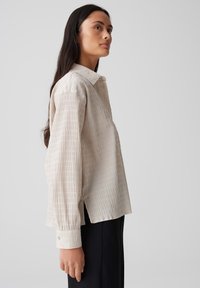Helle beige Bluse mit Kragen und einem strukturierten gestreiften Muster, langen Ärmeln und geknöpften Manschetten; lässiger Schnitt mit seitlichen Schlitzen für mehr Bewegungsfreiheit.