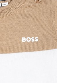 Pull en tricot avec une partie supérieure beige et une partie inférieure blanche. Il présente un col côtelé et le nom de la marque "BOSS" imprimé en blanc.
