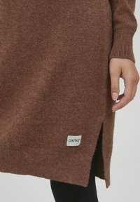 Bruine gebreide trui met geribbelde manchetten en een zijsplit. Heeft een zichtbare stoftextuur en een wit label met het "oxmo"-merk.