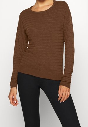 Femme portant un pull côtelé marron ajusté à manches longues et un pantalon noir, debout avec une main légèrement pliée et l’autre détendue.