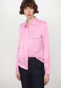 Pink, glossy button-up shirt with a snake-print texture, featuring two chest pockets and a classic collar. Worn with dark blue jeans.
