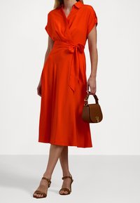 Kvinna klädd i en ljusorange omslagsklänning med korta ärmar och håller en liten brun handväska, i kombination med bruna klacksandaler.